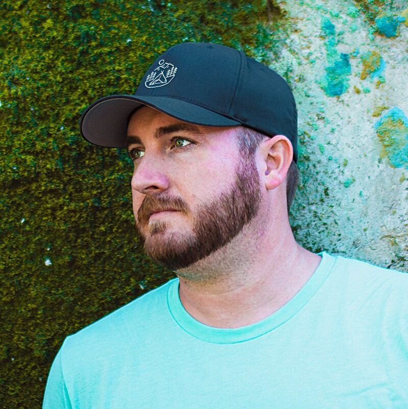A bearded man in a black baseball cap and light blue shirt looks off to the side while standing in front of a mossy, textured wall.