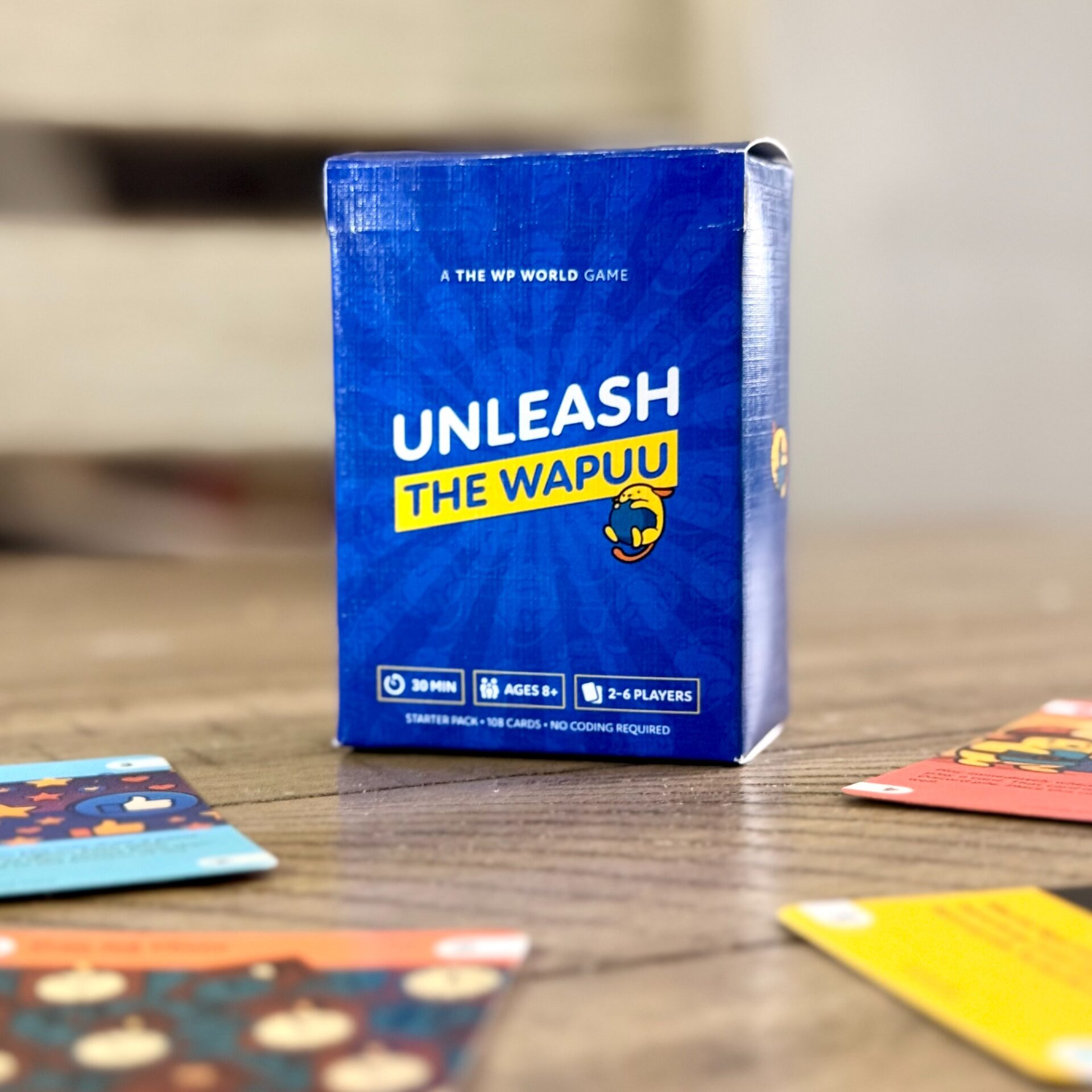 A blue game box labeled “Unleash the Wapuu” sits on a wooden table, surrounded by colorful game cards. The box shows icons for 30 minutes, ages 18+, and 2–6 players.