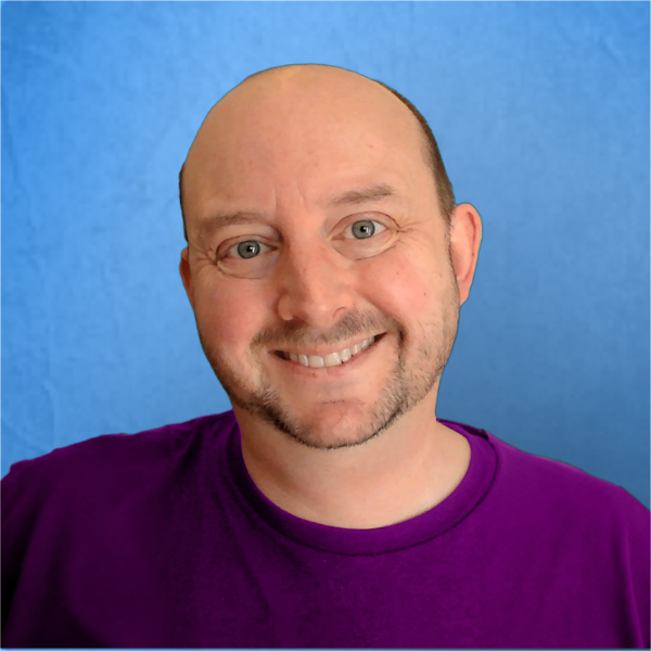 Corey Maass, a smiling man with a bald head and close-cropped beard wearing a purple shirt stands in front of a blue background.