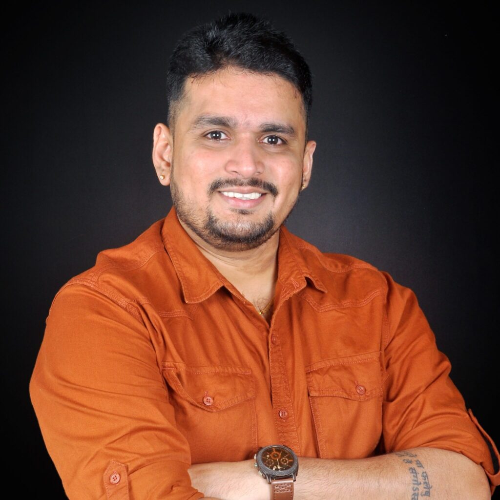 A man with short dark hair and a beard smiles with arms crossed, wearing an orange button-up shirt and a wristwatch, standing against a plain black background.