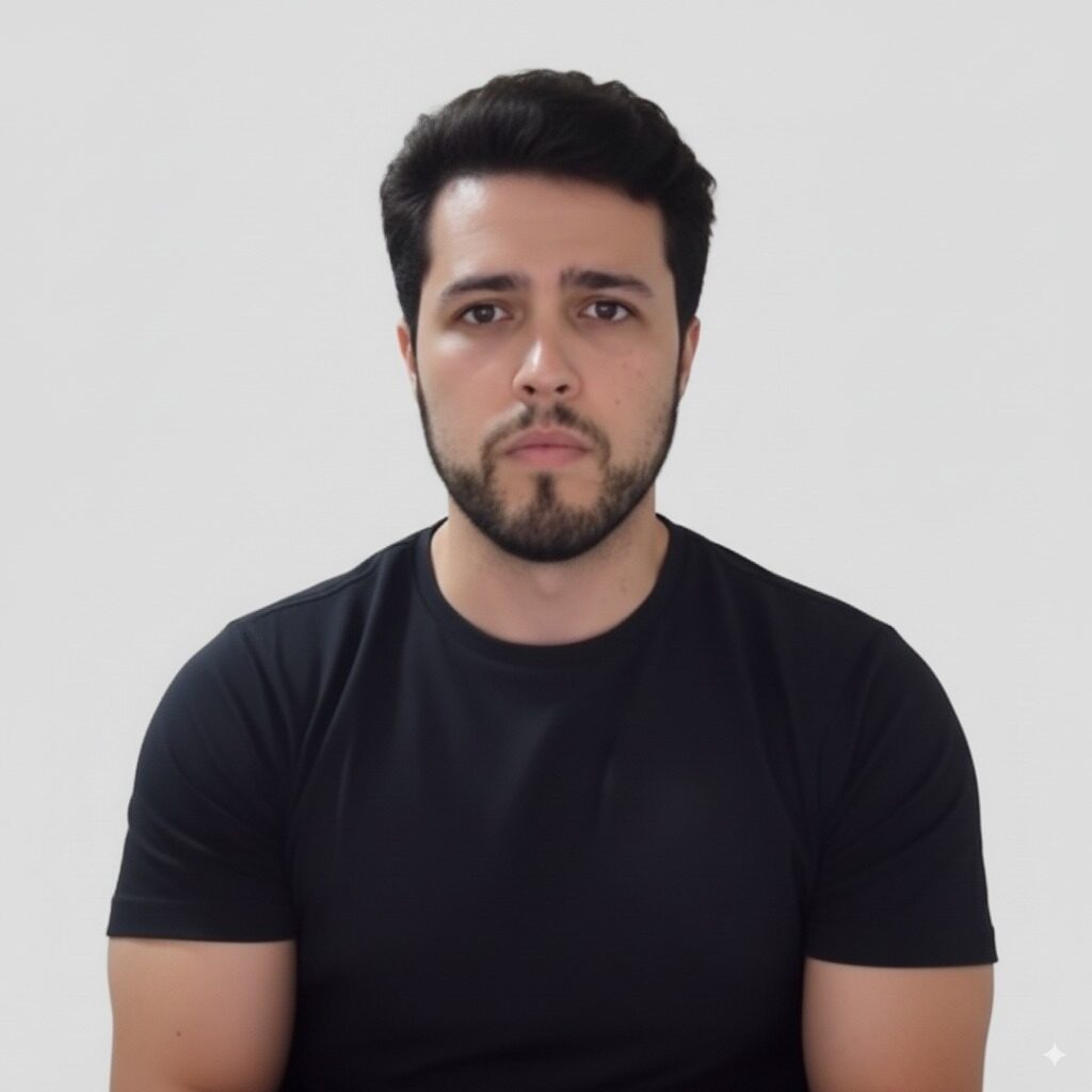A man with short dark hair and a beard, wearing a black t-shirt, sits against a plain light gray background, looking directly at the camera with a neutral expression.