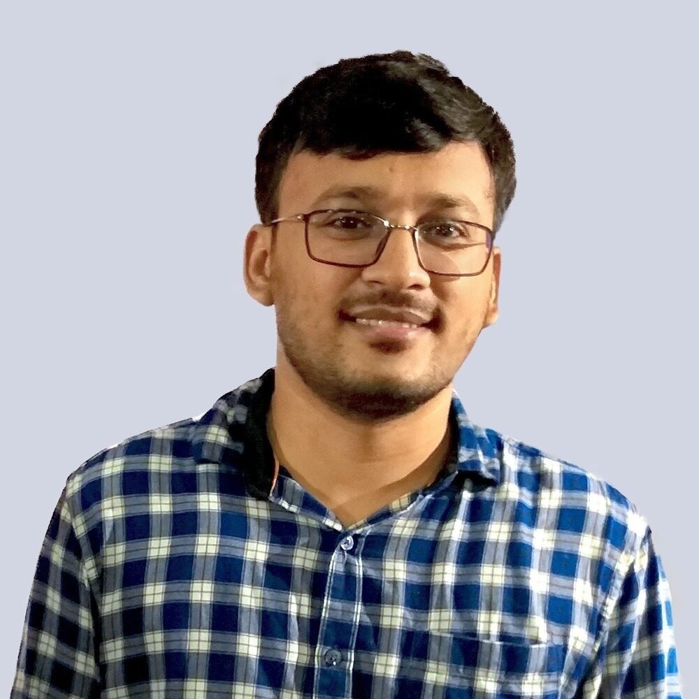 A man with short dark hair, glasses, and a trimmed beard is wearing a blue and white checkered shirt, smiling, and standing against a plain light gray background.