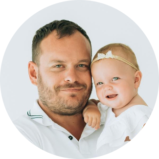 A smiling man with short brown hair and a beard holds a happy baby girl wearing a white outfit and a headband with a bow. Both are looking at the camera against a plain, light background.